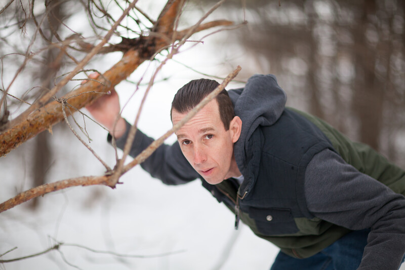 Man leaning toward bare branches in snowy forest, wearing dark jacket and cap.