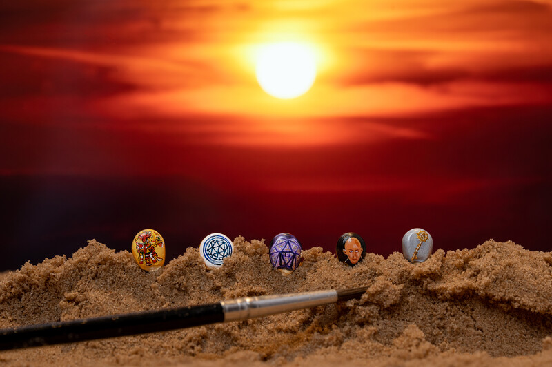 Six decorated coins arranged in a line on sand with a paintbrush, set against a dramatic sunset sky.