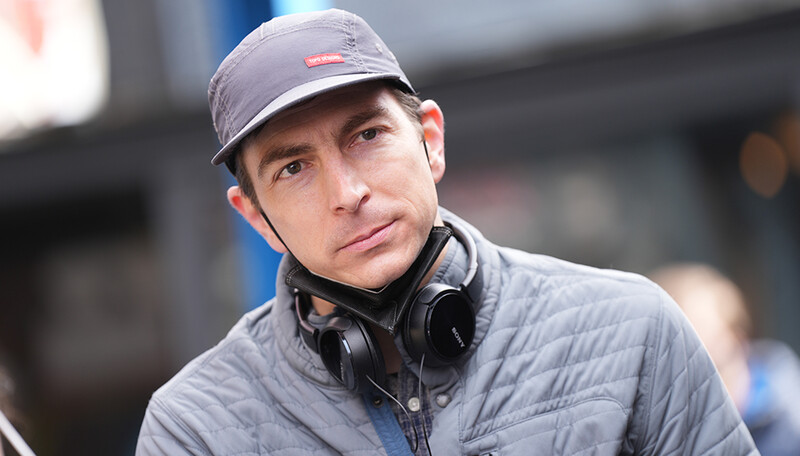 Man wearing a gray quilted jacket and dark cap with headphones around his neck, looking directly at camera.