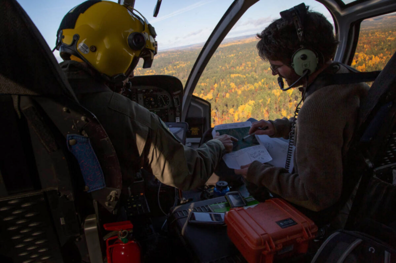 Two people in a helicopter cockpit reviewing documents during flight over forested terrain.