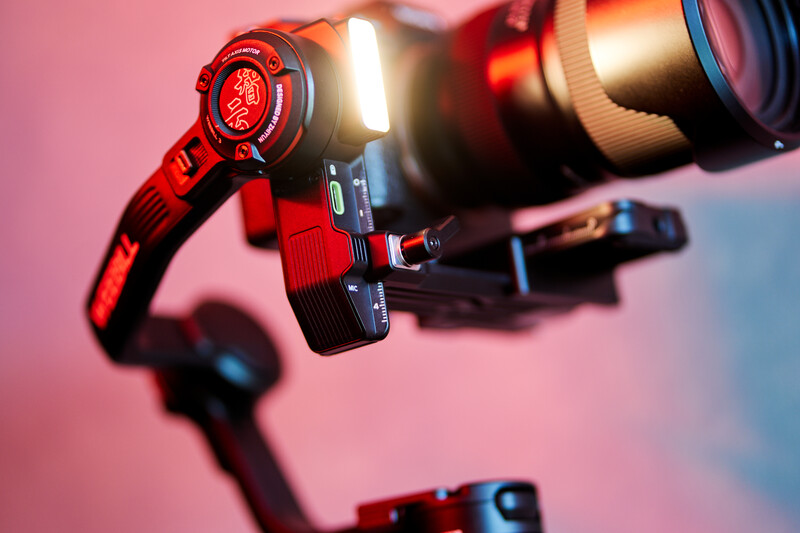 Red camera rig with focus motor and lens mounted on pink and blue gradient background.