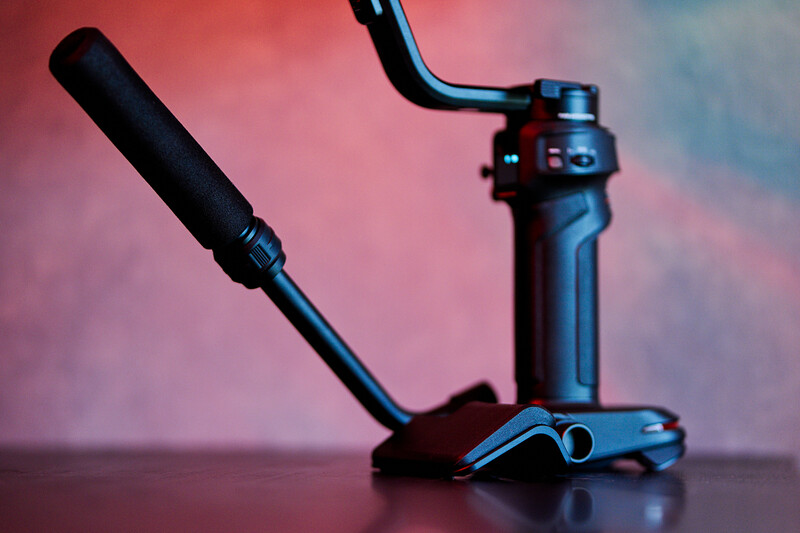 Professional camera gimbal with mounted microphone against colorful pink and blue gradient background.