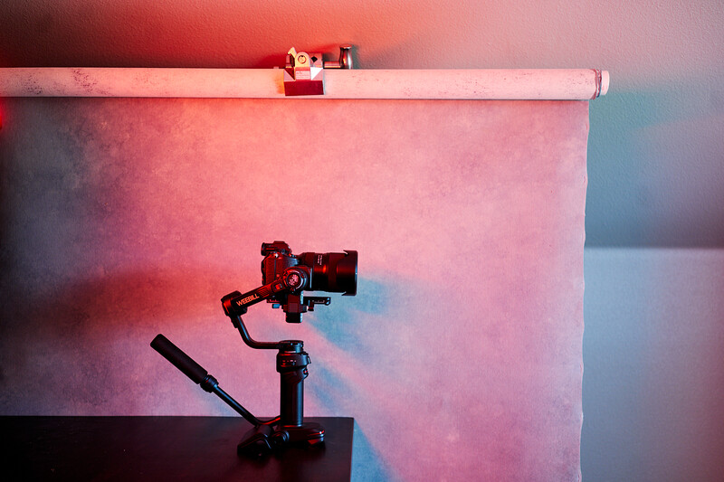 Red gimbal-mounted camera on tripod against pink and blue gradient backdrop with studio lighting.