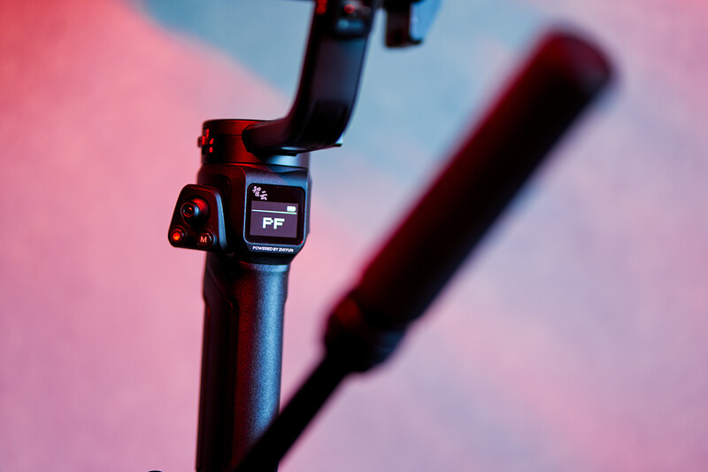 Close-up of a camera gimbal's pan-tilt head with control display against a pink and blue gradient background.