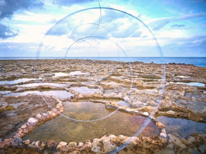 Tide pool with concentric stone circles on rocky coastal shoreline under bright sky.