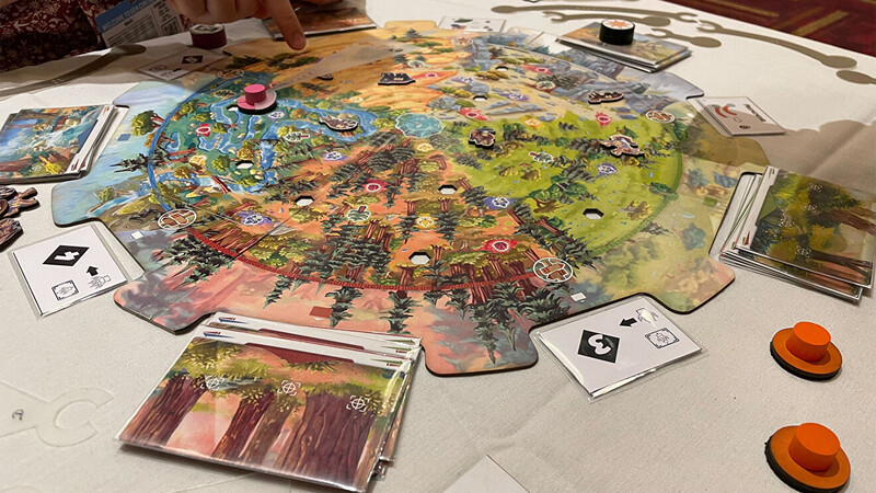 Board game components spread across a table during gameplay, including a colorful illustrated game board, player tokens, cards, and orange player pieces.