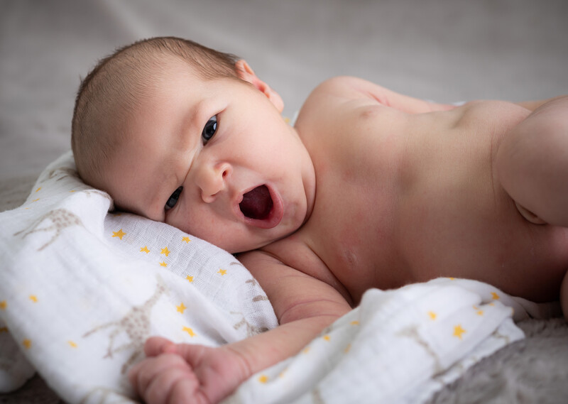 Newborn baby lying on white bedding with yellow stars, looking at camera with open mouth.