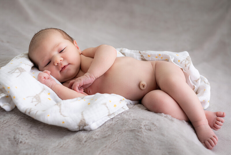 Newborn baby sleeping peacefully on a white patterned blanket against a gray backdrop.
