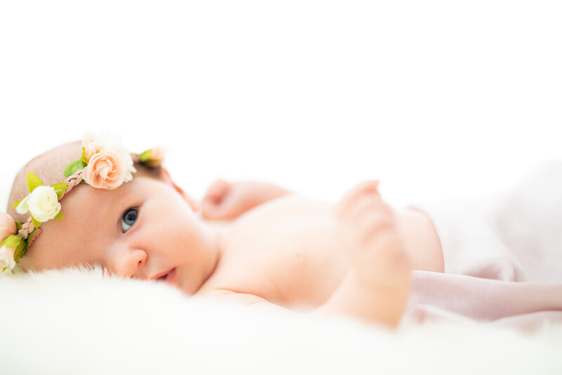 Newborn baby wearing a floral crown, lying on white bedding and looking toward camera.