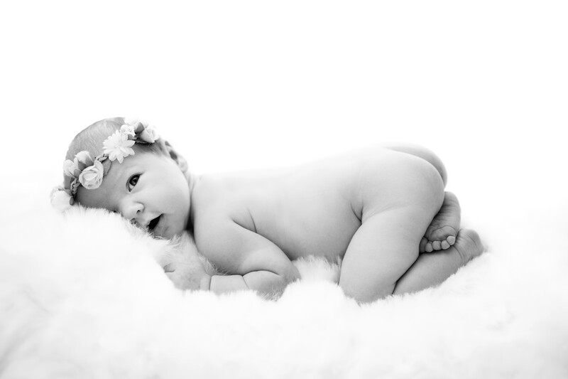 Newborn infant in fetal position on white fluffy fabric, wearing delicate floral headpiece.