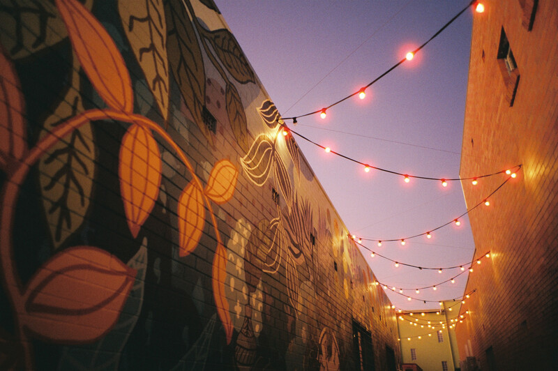 Warm string lights strung between two buildings decorated with large-scale mural artwork at dusk.