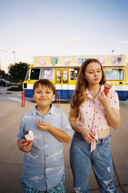 Two children standing outdoors in front of a blue and yellow food truck during golden hour.