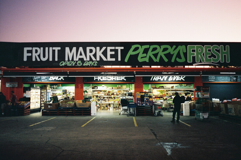 Storefront of a fruit market with illuminated signage at dusk, featuring red and dark facade with multiple shop windows.