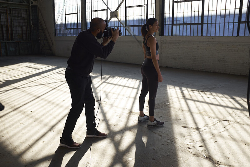 Photographer in dark clothing shooting a portrait of a woman in athletic wear on a bright rooftop with blue water visible in the background.