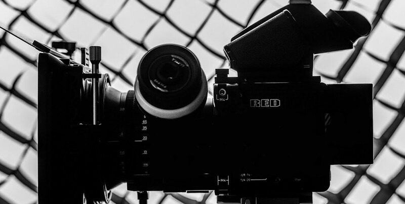 High-contrast silhouette of a RED cinema camera with lens and monitoring equipment against a geometric ceiling grid.