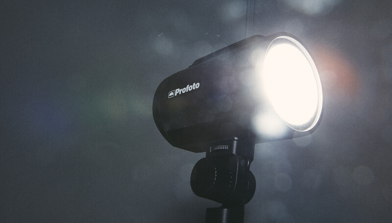 Profoto studio strobe mounted on stand, illuminated against dark cloudy sky with dramatic light spill.