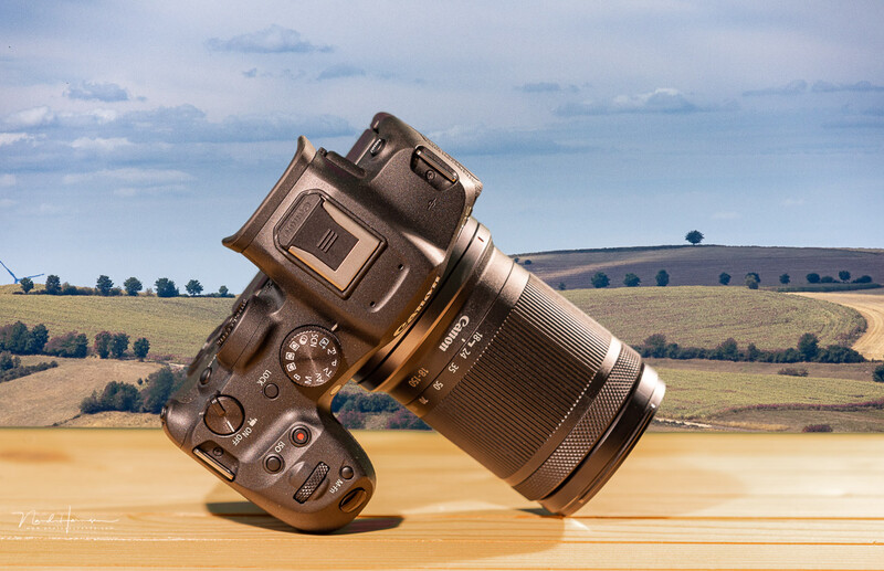 DSLR camera with attached lens tilted at an angle on a wooden surface, with a rural landscape in the background.