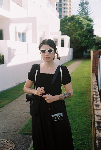 Woman in black outfit and sunglasses standing on a residential pathway with modern white building and green lawn.
