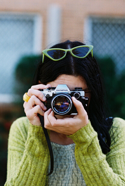 Photographer holding a vintage rangefinder camera up to their face with yellow-framed sunglasses perched on their head.