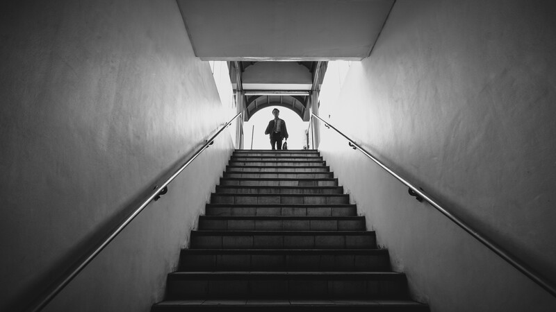 A solitary figure ascending a long staircase toward bright light at the top, photographed from below in black and white.