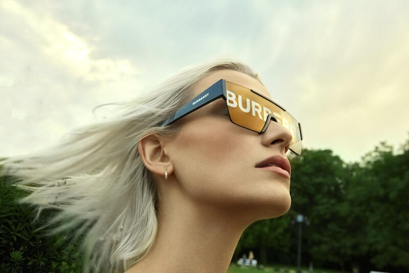 Woman with long platinum blonde hair wearing oversized sunglasses with gold lettering, shot in profile against a cloudy sky with green foliage behind.