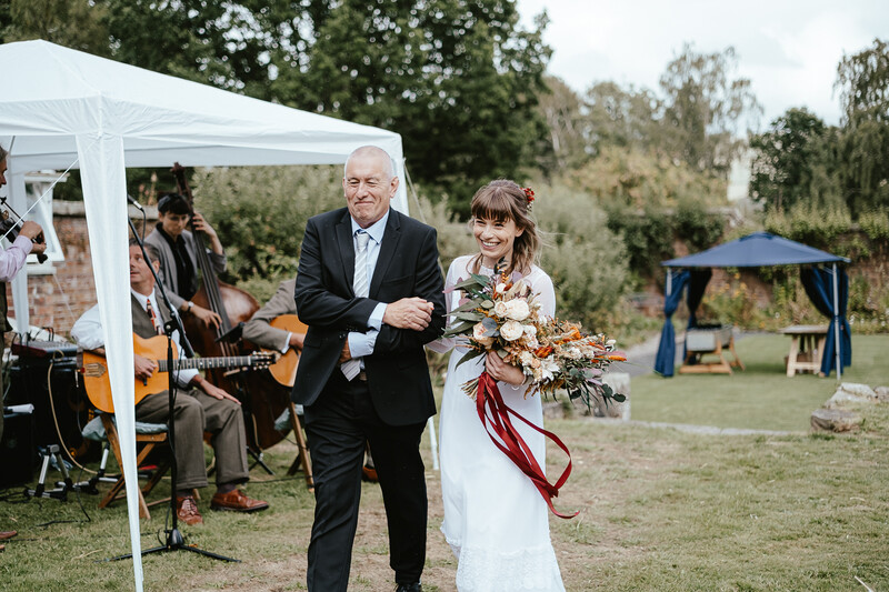 Bride and groom walking together at outdoor wedding ceremony with live musicians and guests in background.