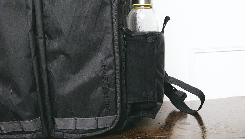 Black camera bag with water bottle pocket displayed on wooden surface against white background.