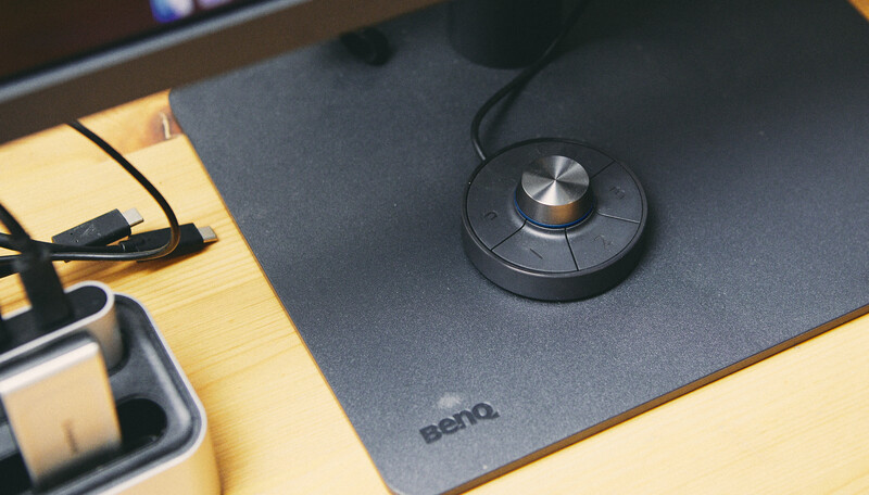 BenQ tablet stylus pen resting on a dark gray graphics tablet with wooden desk surface visible.
