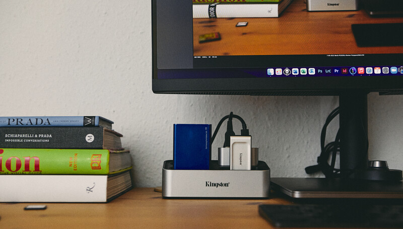 Organized desk workspace with stacked books, monitor displaying desktop, docking station, and headphones on wooden surface.
