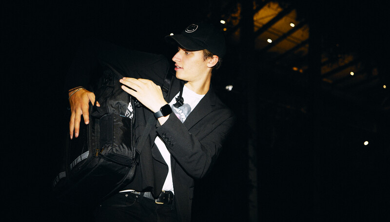 Person in dark blazer and cap holding black camera equipment against dark background with warm overhead lighting.