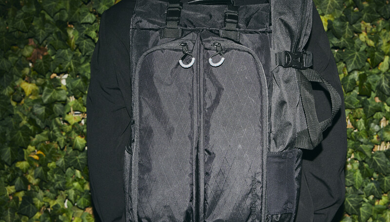 Dark gray camera backpack with dual zipper compartments and metal hardware against green foliage background.