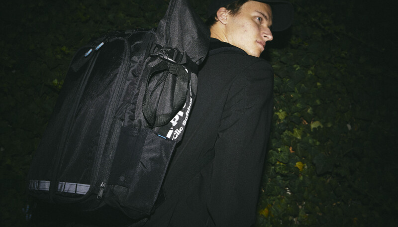 Man wearing black jacket and tactical backpack standing against dark foliage at night.