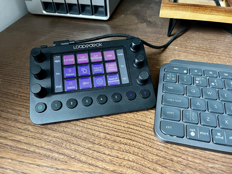 Loupedeck control panel with illuminated purple buttons next to a wireless keyboard on a wooden desk.