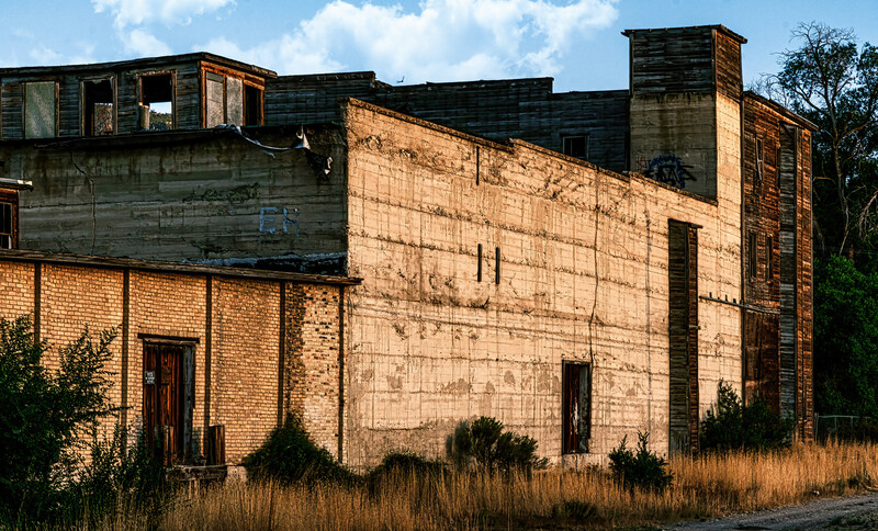 Abandoned brick industrial building with boarded windows and overgrown vegetation in afternoon sunlight.