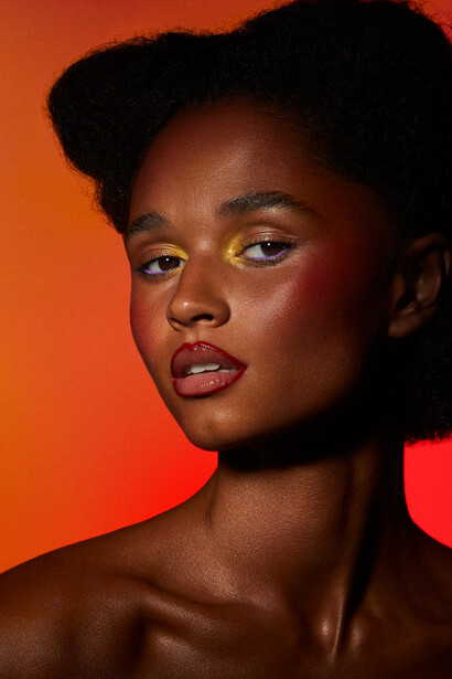 Woman with bold yellow eyeshadow against a warm orange and red gradient background.