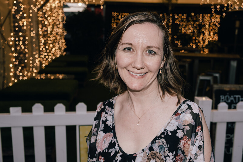 Woman smiling at camera wearing floral print top, warm string lights blurred in background.