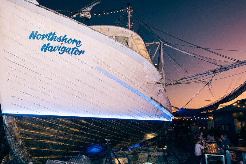Illuminated sailing vessel with blue accent lighting at dusk, moored alongside a pier with event space.