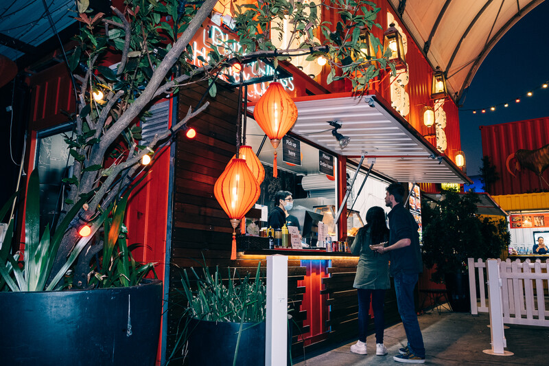 Interior of an Asian-style bar or restaurant with hanging lanterns, potted plants, and customers at the counter.