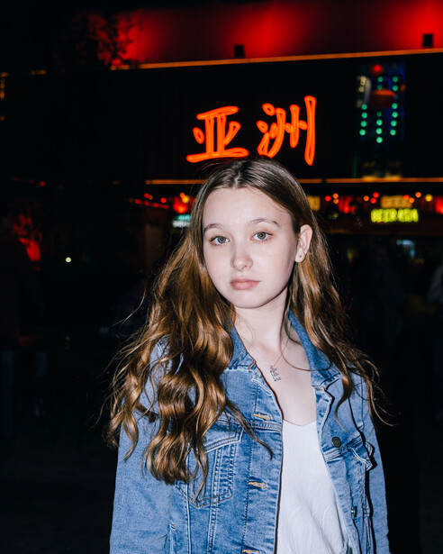 Young woman with long wavy hair wearing a light blue denim shirt stands against neon signage at night.