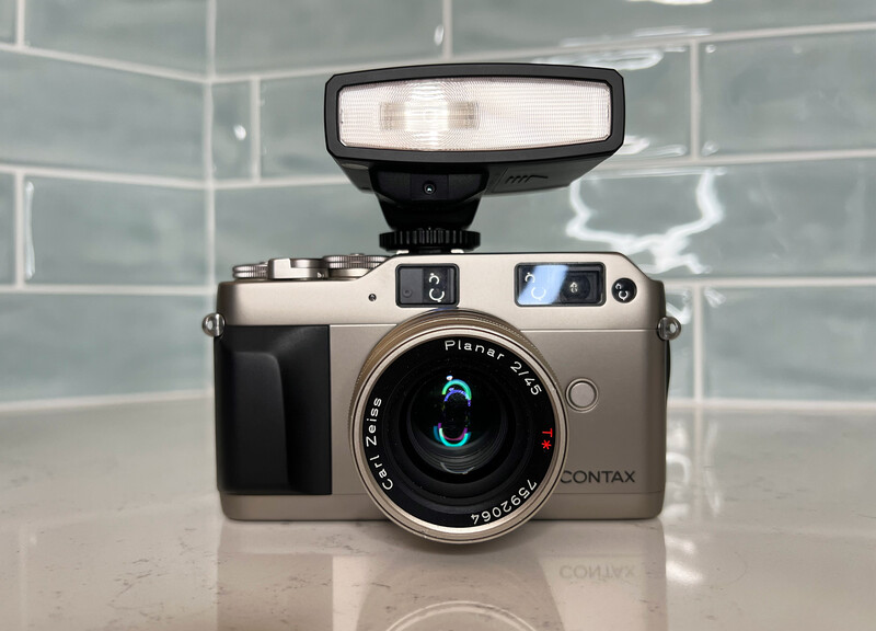 Contax rangefinder camera with mounted flash against subway tile background.