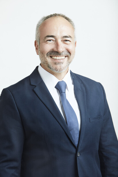 Professional headshot of a man in dark business suit and blue tie smiling at camera.