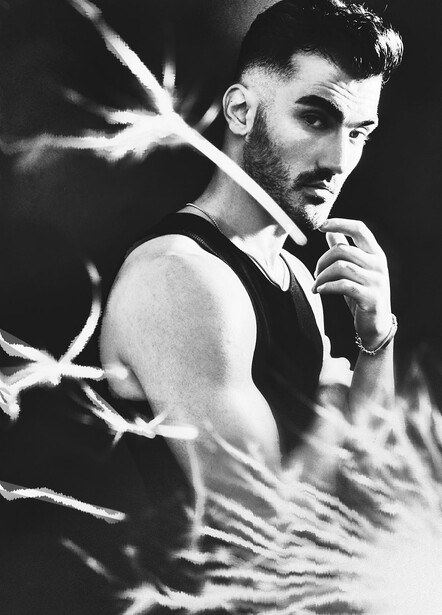 Bearded man in sleeveless top posed against branches in black and white portrait.