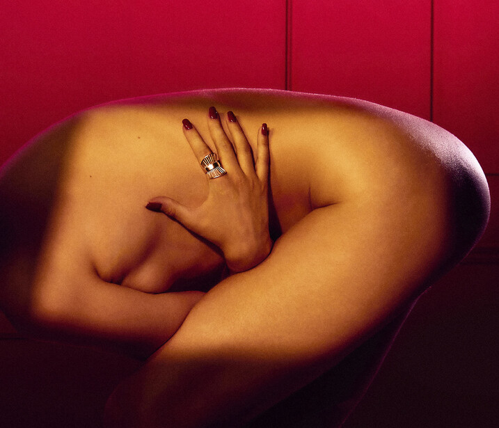 Woman's arm and hand with manicured nails resting against her body against a deep red backdrop.