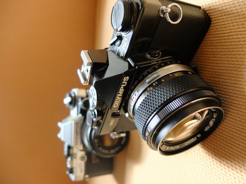 Two vintage Olympus film cameras displayed on a tan textured surface, with a black camera in sharp focus in the foreground.