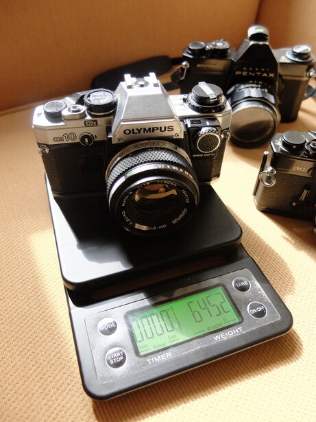 Olympus film camera on a digital scale showing its weight, surrounded by other vintage camera equipment.