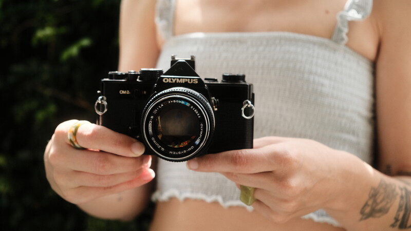 Person in light gray tank top holding a black Olympus OM-2 film camera with 50mm lens.