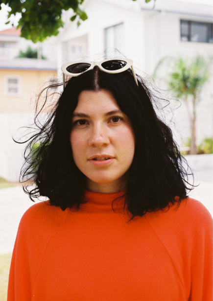 Woman with shoulder-length dark hair wearing a bright orange crew-neck shirt and white oval sunglasses pushed up on her head.