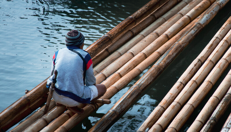 Person sitting on bamboo raft logs floating in turquoise water.