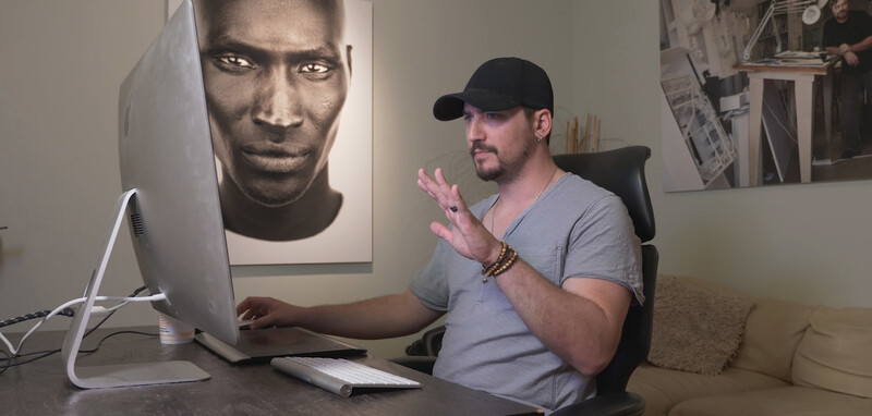 Photographer gesturing while discussing work at desk with monitor displaying portrait photography.