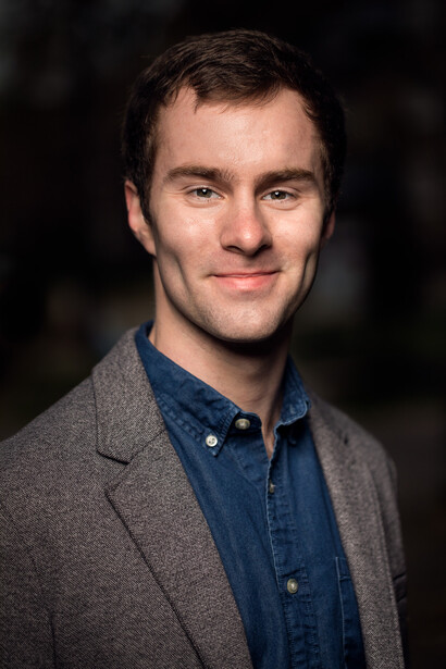 Professional portrait of a man wearing a gray cardigan over a denim shirt against a dark background.